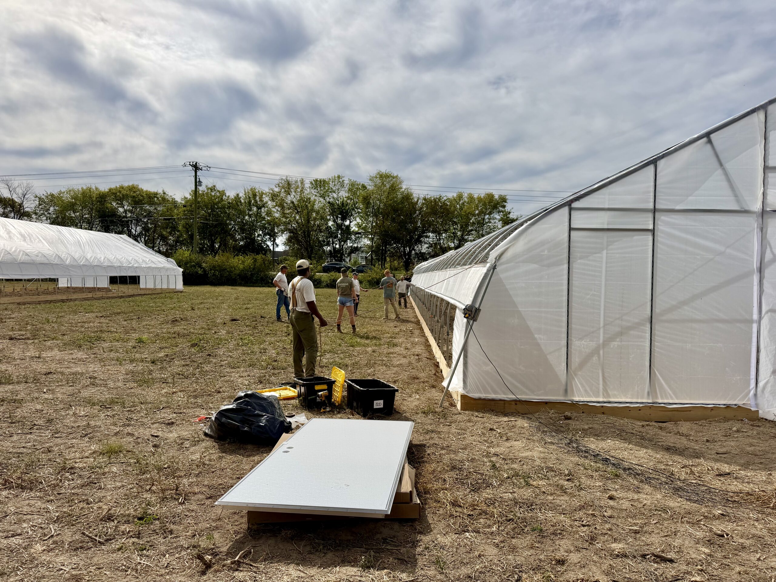Workers stand beside a newly covered hoop house with tools on the ground.